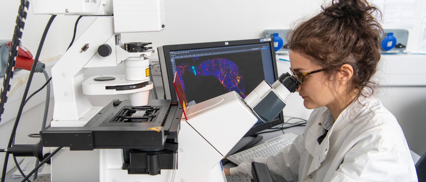 Female student looking through a microscope in a lab with a computer screen displaying visuals