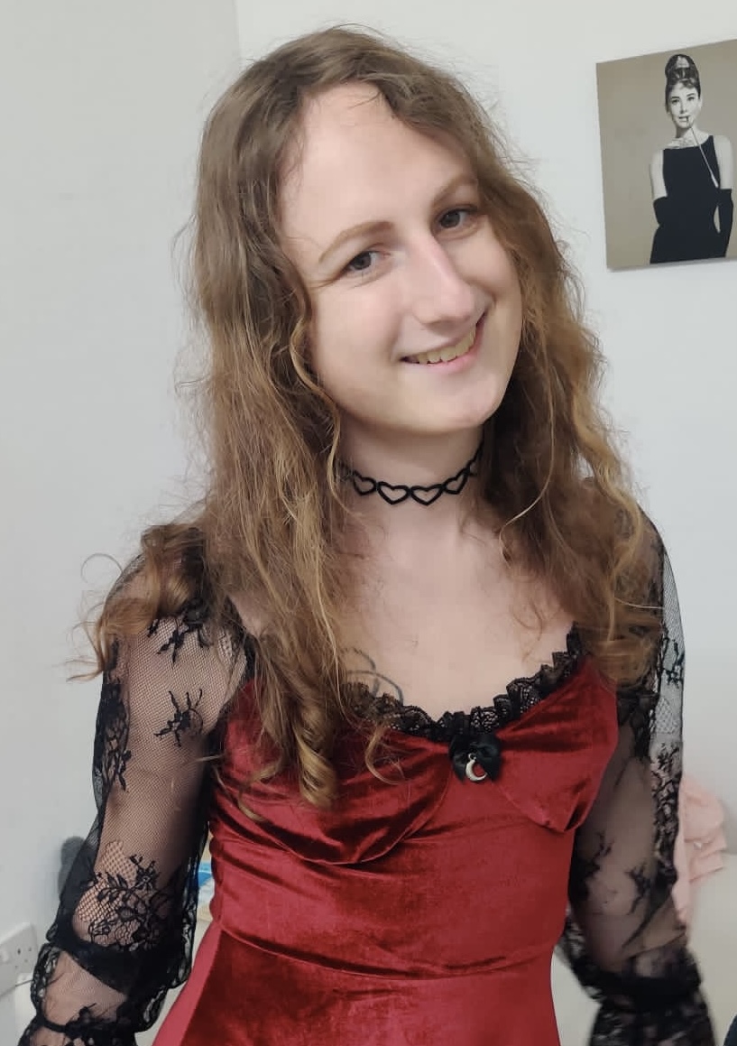 Samantha Pilgrim in a red and black Gothic dress.  She is smiling and wearing a necklace with hearts on it.  A portrait of Audrey Hepburn hangs in the background.  