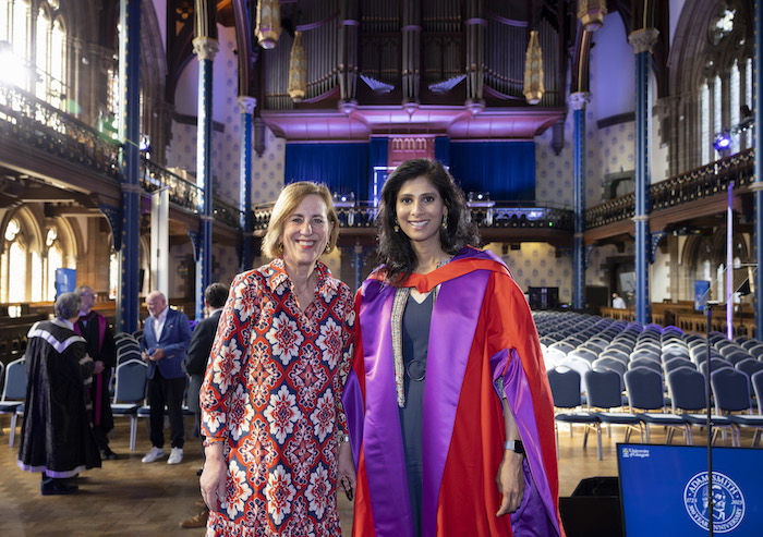 Kirsty Wark and Gita Gopinath stand in the Victorian era Bute Hall before the lecture so there are rows of seats behind them. Both are smiling and Gita Gopinath is wearing red and purple academic robes 