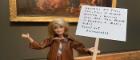 blonde barbie doll holding placard in front of painting