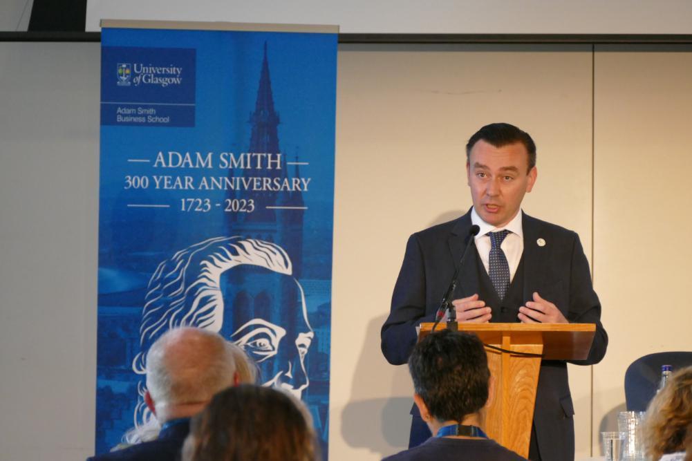 Craig Smith speaking at a podium with the Adam Smith 300 banner with the university of Glasgow logo and text stating Adam Smith 300 year anniversary 1723 - 2023 Source: ASBS
