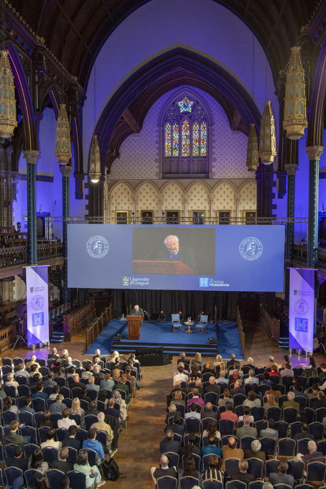 A view of the Bute hall with an audience siting listening to Angus Deaton. Behind him is a large screen with a live recording of him with the Adam Smith tercentenary logo and the University of Glasgow and Hunter Foundation logo. Source: Charlotte Morris, External Relations