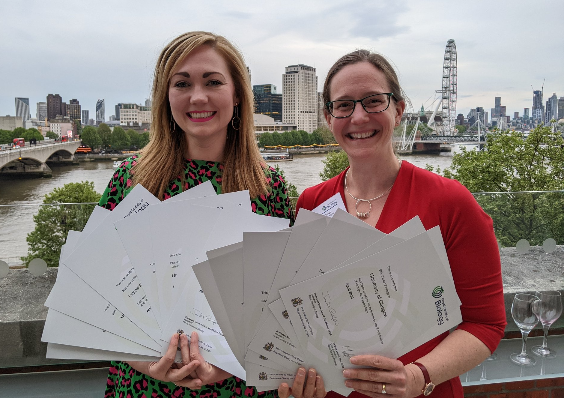 Dr Laura McCaughey and Dr Anna McGregor stood holding paper certificates fanned out with London behind them