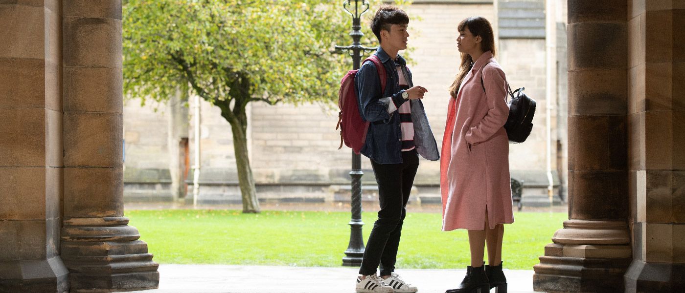 Two students chatting together with the University buildings in background