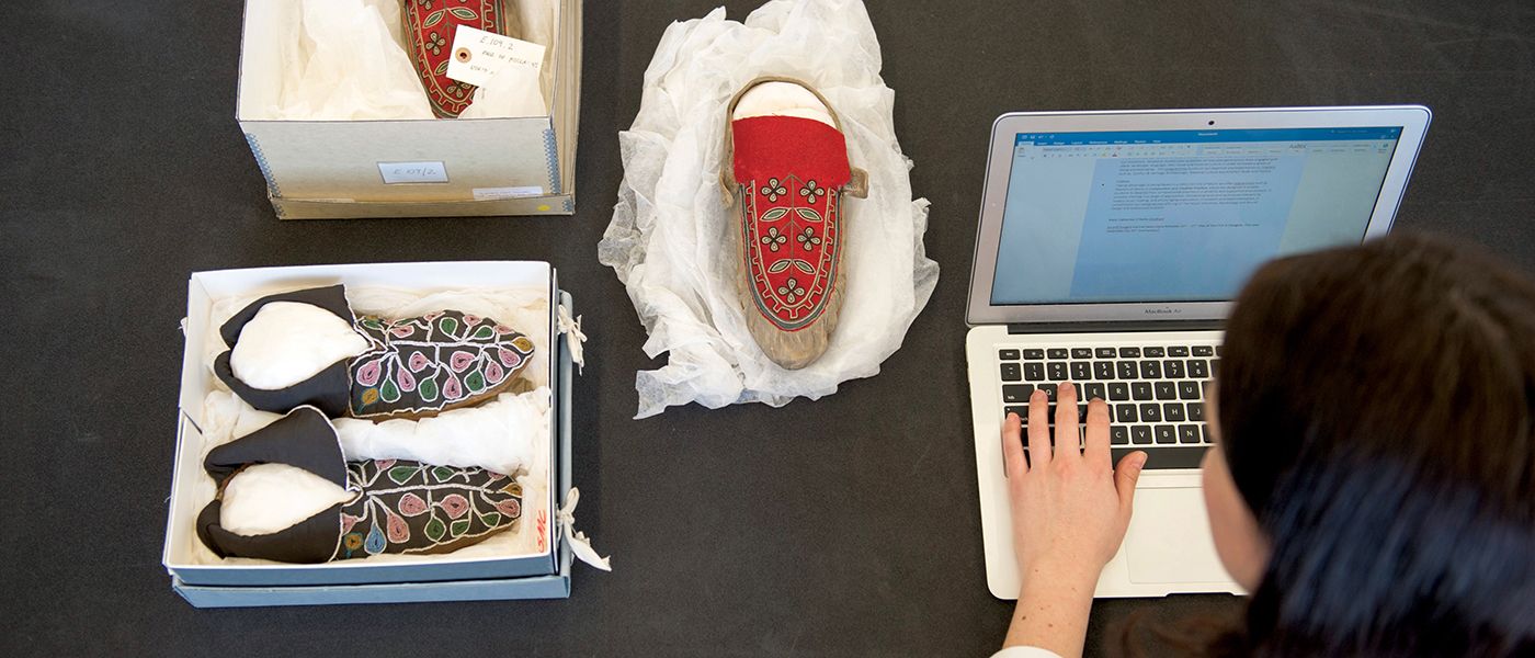 A woman writes on a laptop while studying elaborately decorated shoes from a museum collection