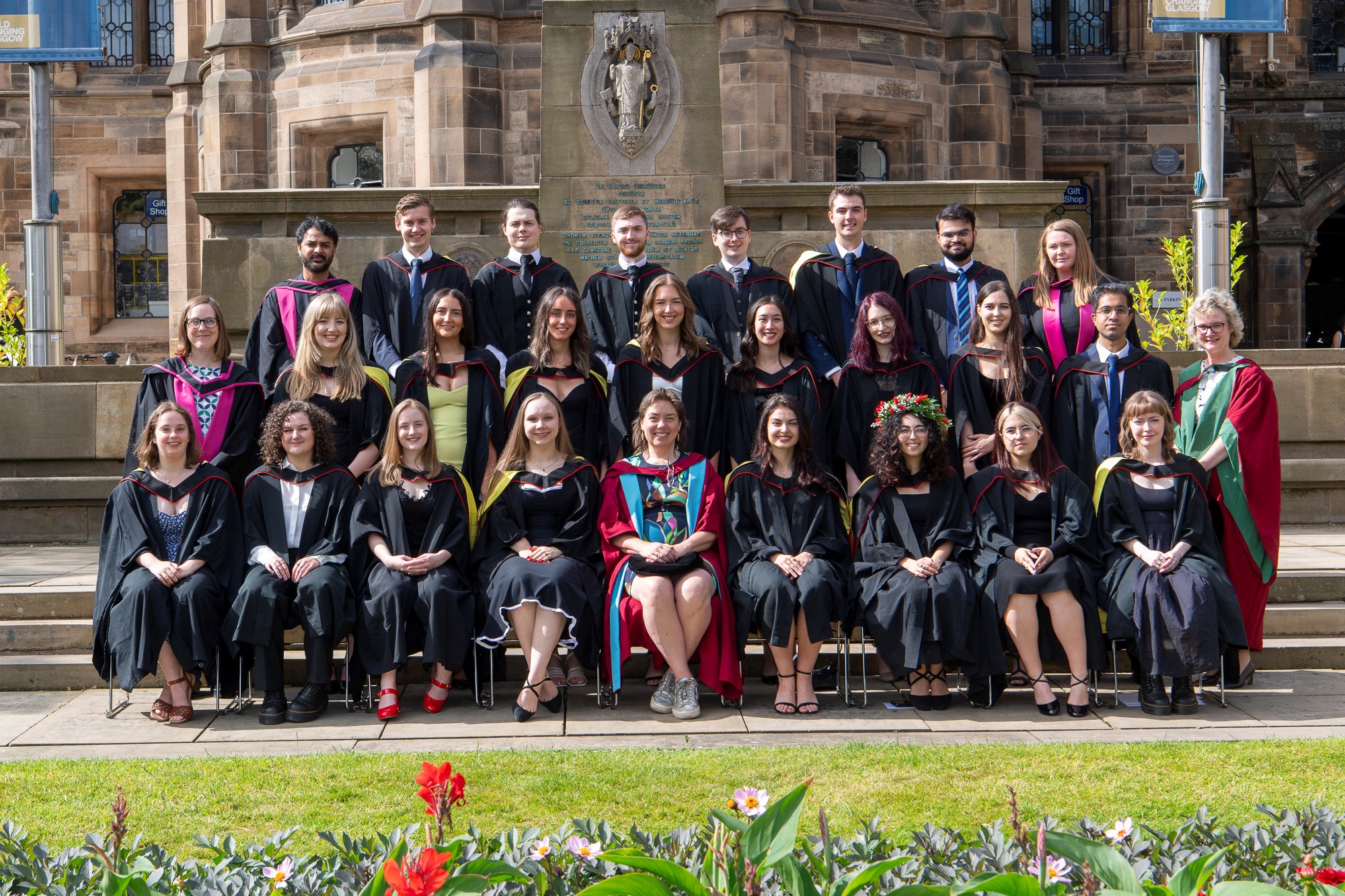 A mix of students and academics sat and stood in three rows outside in graduation attire outside the UofG main building