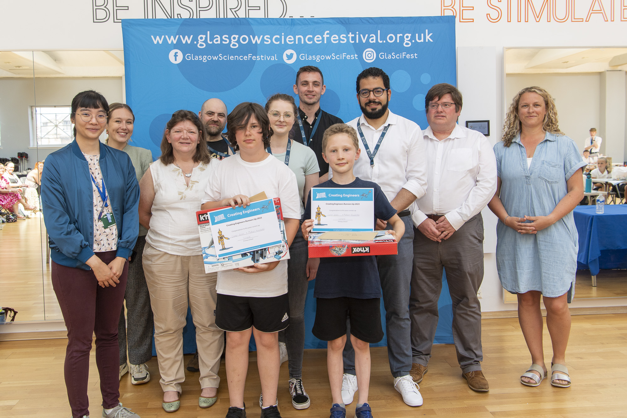Photograph showing the runners up holding their prizes. Behind them are the eight judges and the Glasgow Science Festival director.