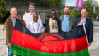 Prof Mike Barrett, Prof Stuart Gray, Malombo Kayira, MacPherson Mbeya stood together on campus holding a Malawi flag in front of them