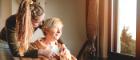 Image of two woman in front of a window, one sitting on a chair and the other helps to put a shawl around her shoulders. Credit Macmillan Cancer Support